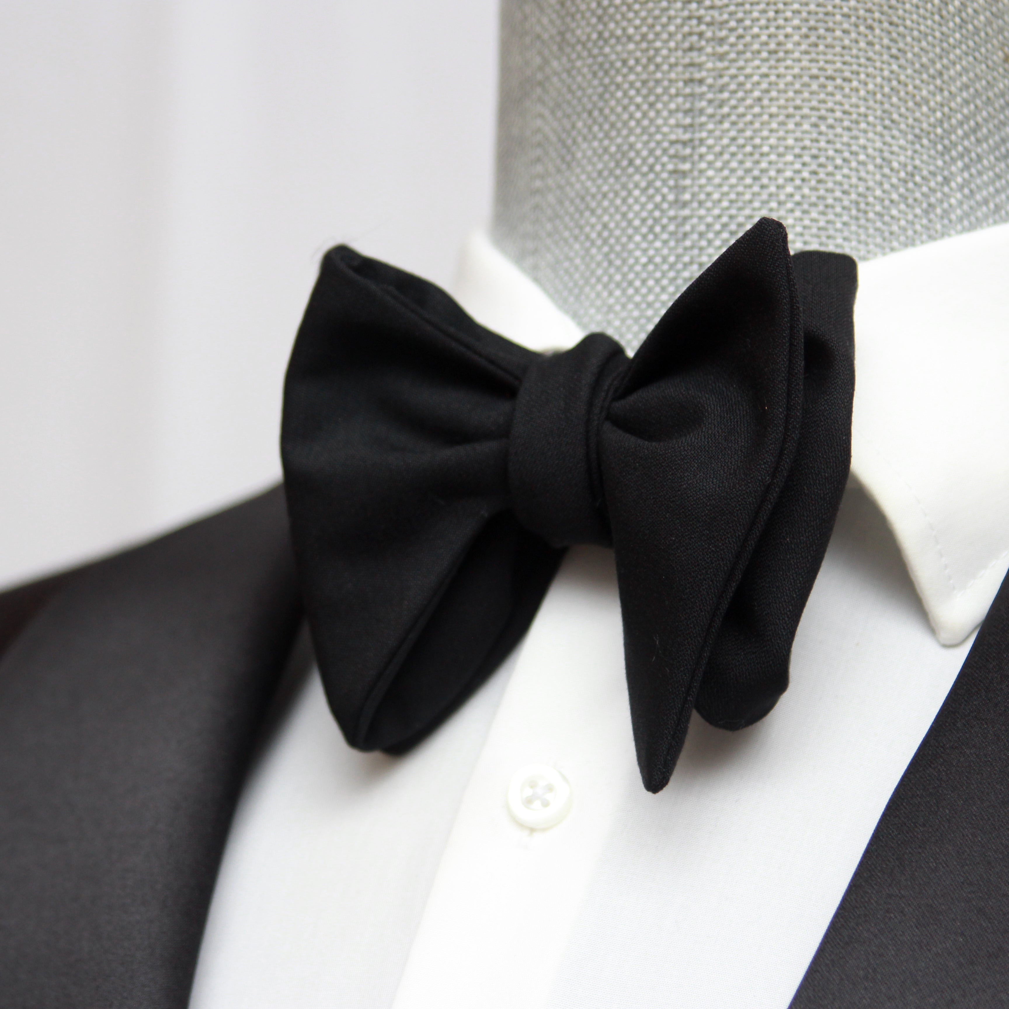 A black wool bow tie displayed on a mannequin wearing a black tuxedo.