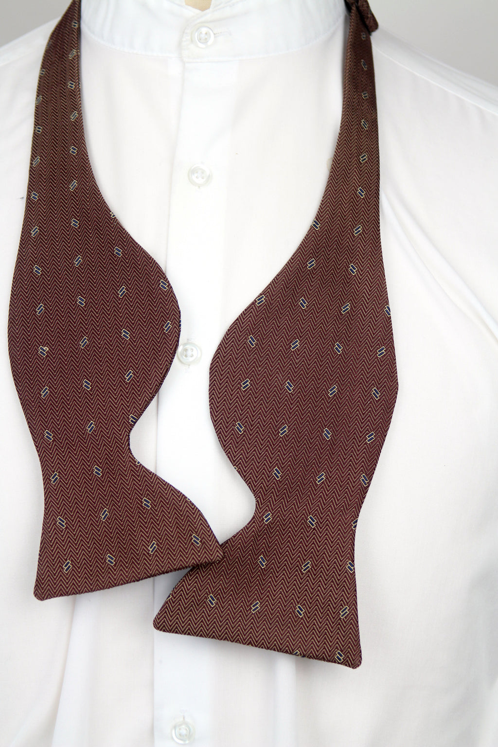 A shape bow tie in a dark brown color with a small repeating paisley pattern, styled on a white collared dress shirt.