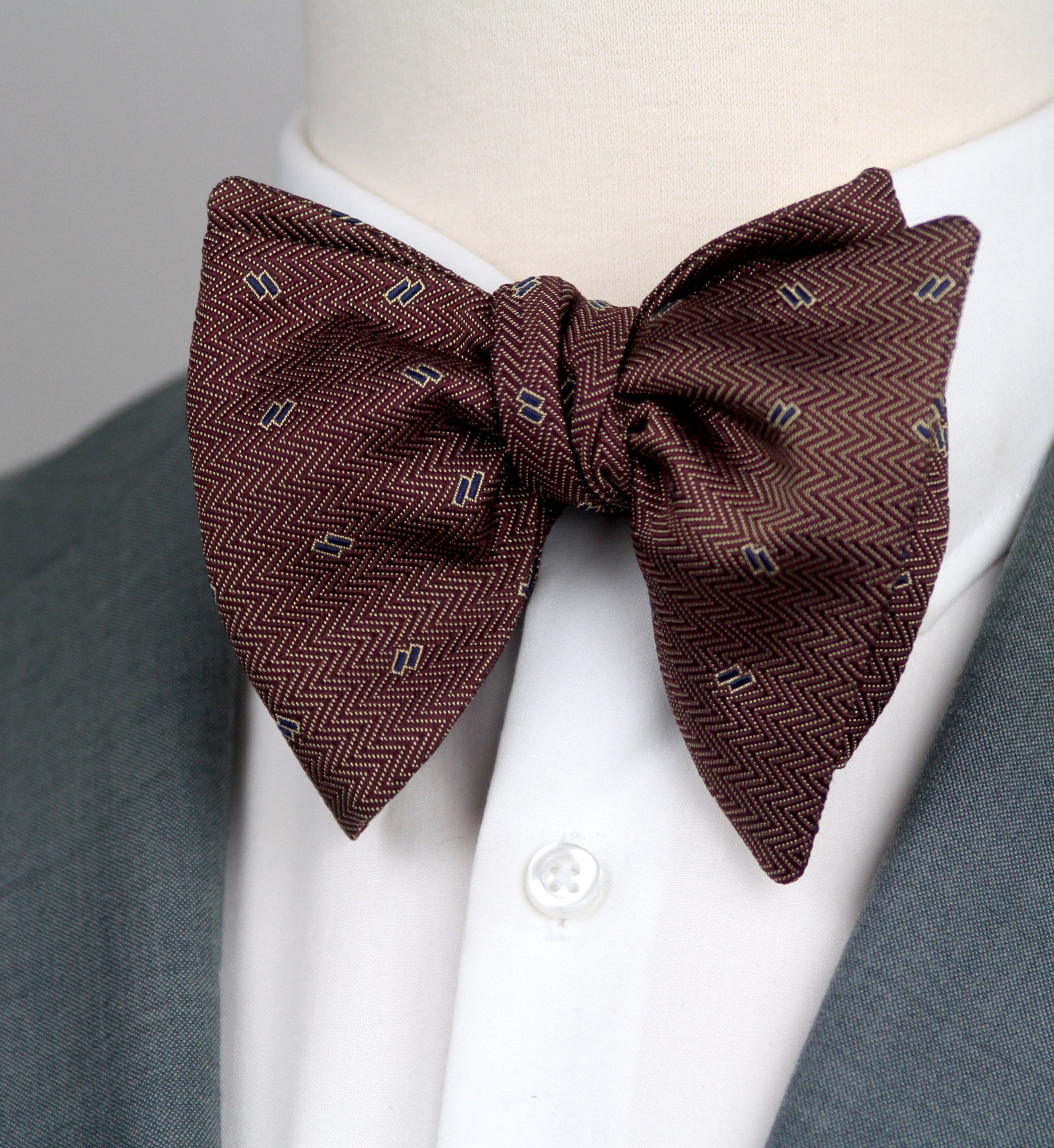 A pre-tied bow tie with a brown and burgundy herringbone pattern and small white geometric accents, styled on a white dress shirt collar and gray suit jacket.