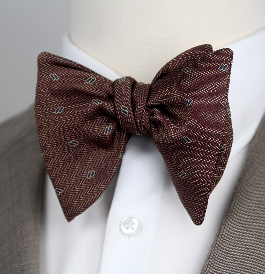 A pre-tied bow tie in a dark brown herringbone pattern with small light blue and white geometric accents, styled on a white dress shirt collar and a gray suit jacket.