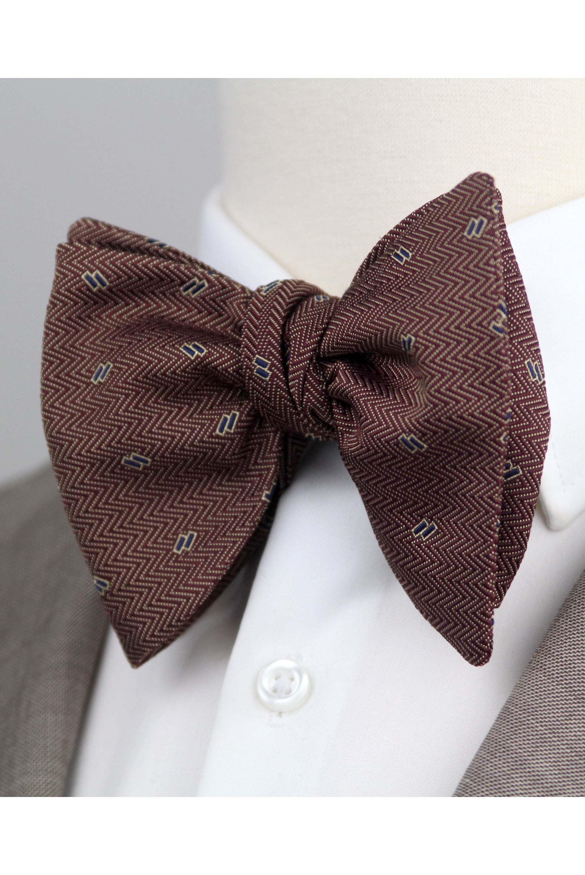 A brown pre-tied bow tie with a small geometric pattern, displayed on a white dress shirt collar and mannequin neck.