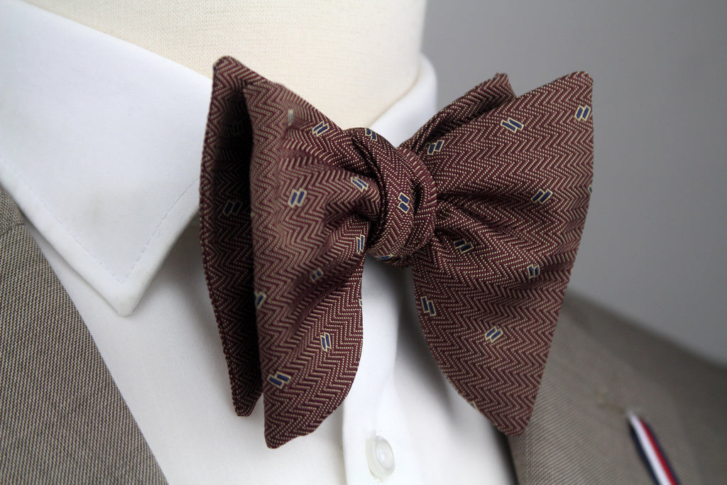 A brown herringbone patterned bow tie with small white geometric accents, styled on a white dress shirt collar and gray suit jacket.