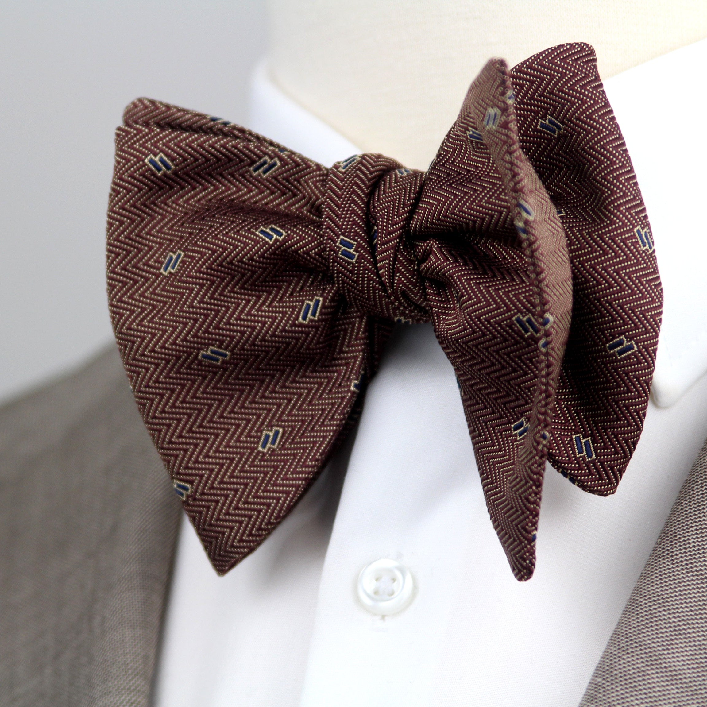 A brown herringbone patterned bow tie with small geometric accents, styled on a white dress shirt collar and gray suit jacket against a light gray background.