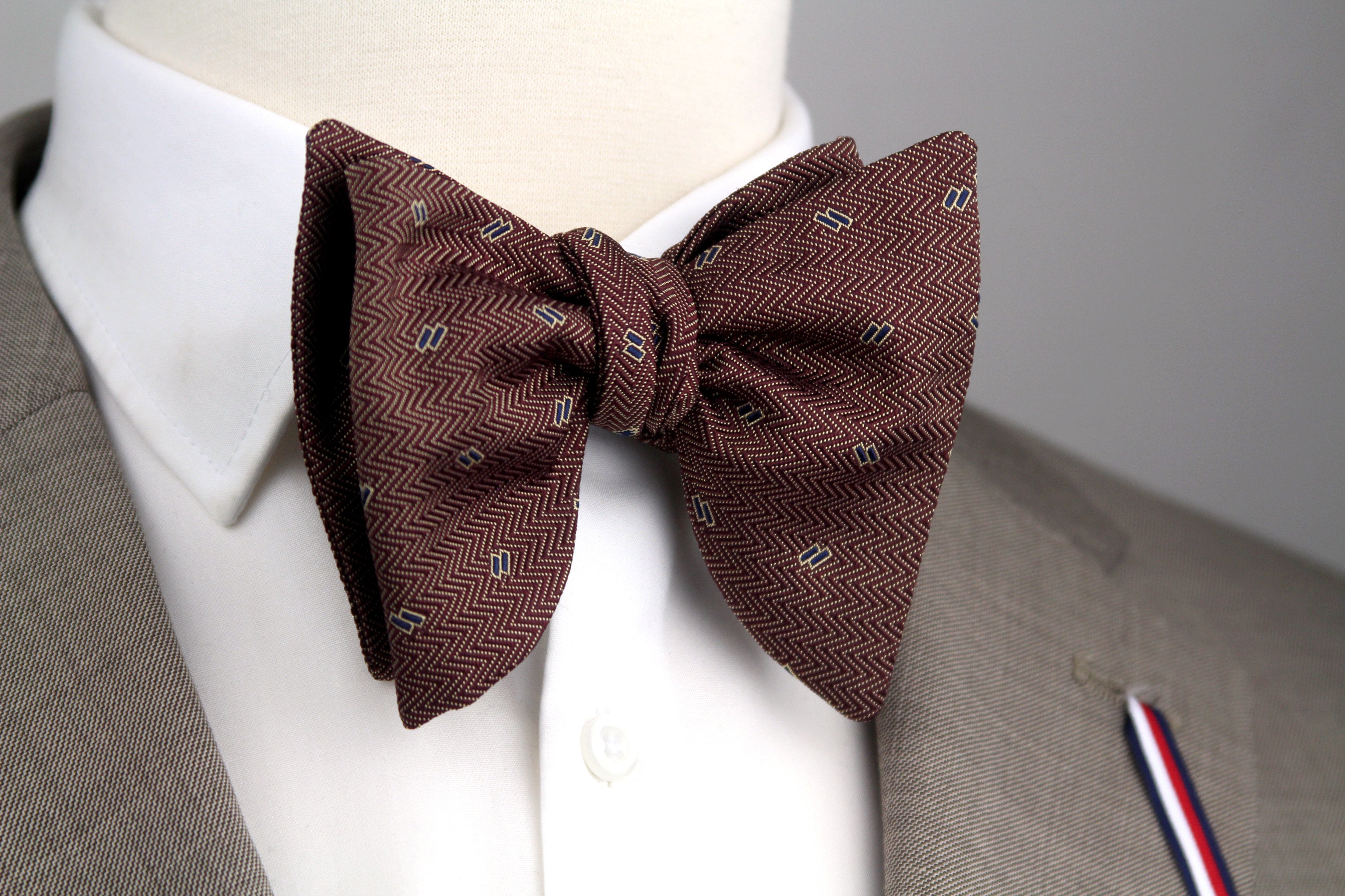 A pre-tied bow tie in dark brown with a fine herringbone pattern and small repeating geometric motifs, styled on a white dress shirt collar and tan suit jacket.