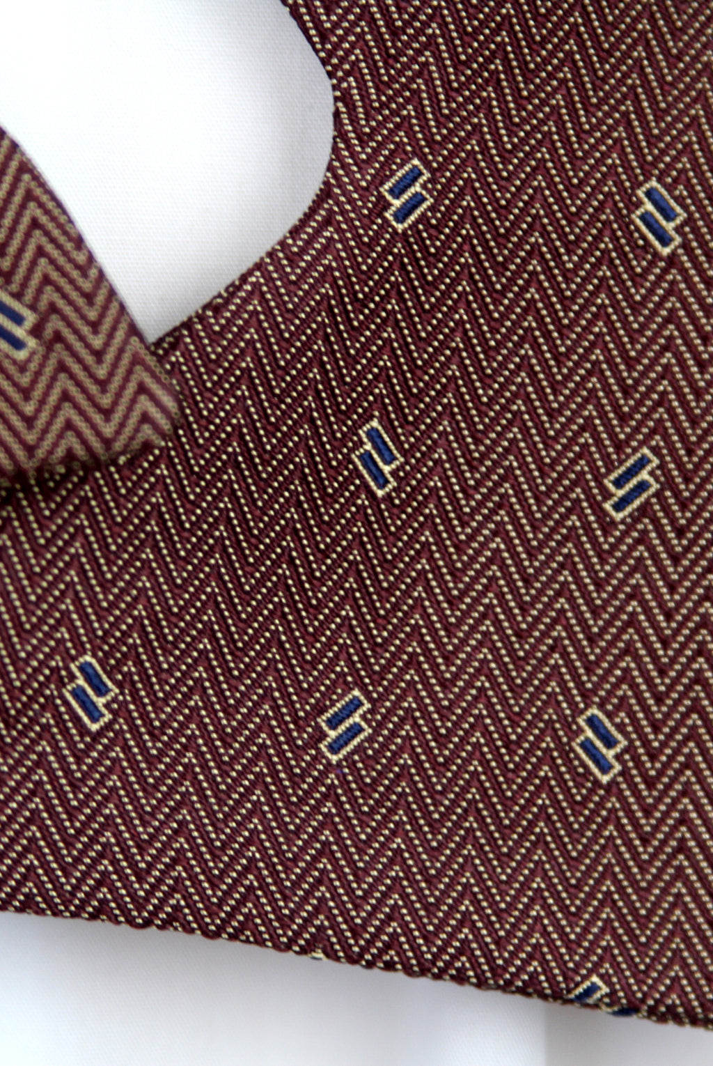 A close-up of a brown and beige herringbone patterned fabric with small blue and gold embroidered letter accents, displayed on a white mannequin torso.