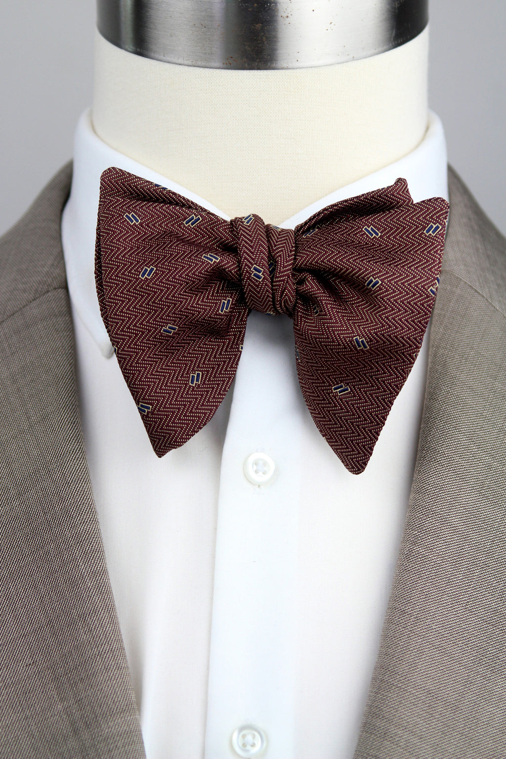 A dark brown herringbone patterned bow tie with small white and gray accents, styled on a white dress shirt and taupe suit jacket displayed on a mannequin torso.