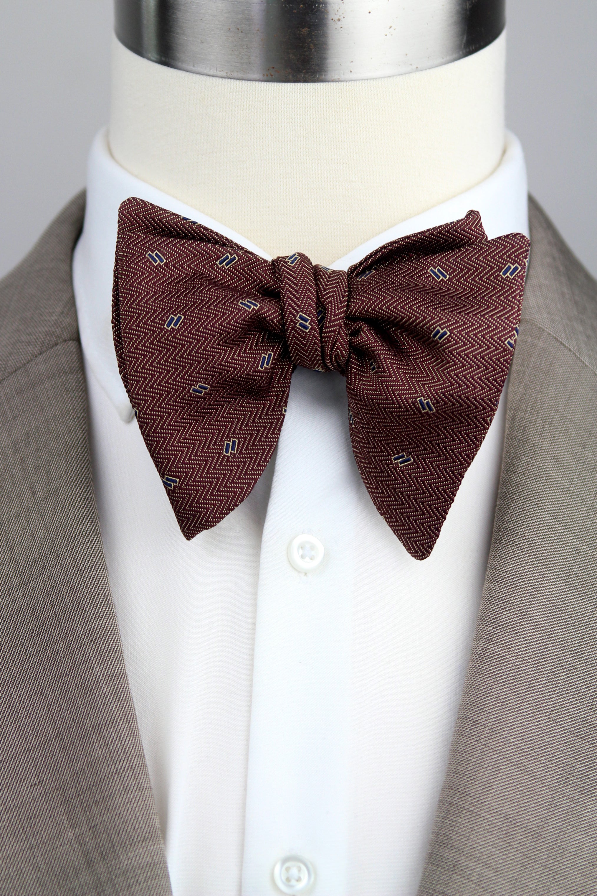 A dark brown herringbone patterned bow tie with small white and gray accents, styled on a white dress shirt and taupe suit jacket displayed on a mannequin torso.