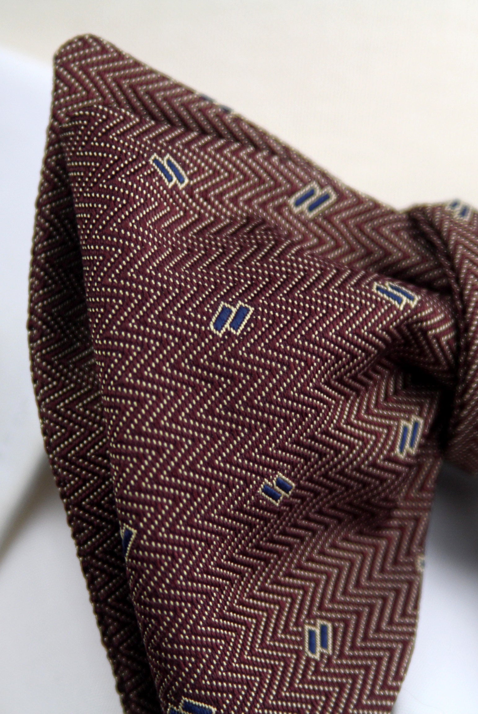 A close-up of a brown herringbone patterned tie with small blue and white embroidered accents, tied in a knot and draped on a white surface.