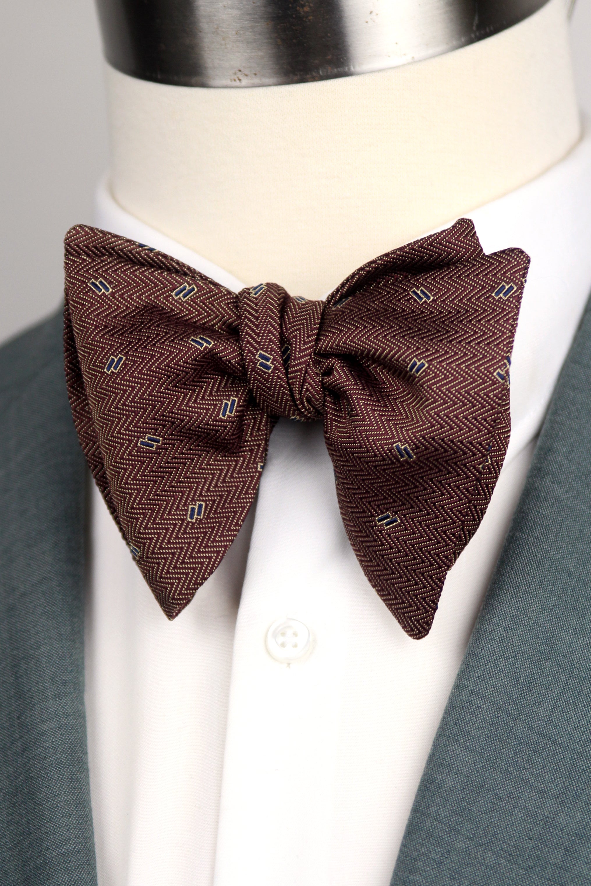 A brown herringbone patterned bow tie with small white and black geometric accents, styled on a mannequin wearing a white dress shirt and gray suit jacket.