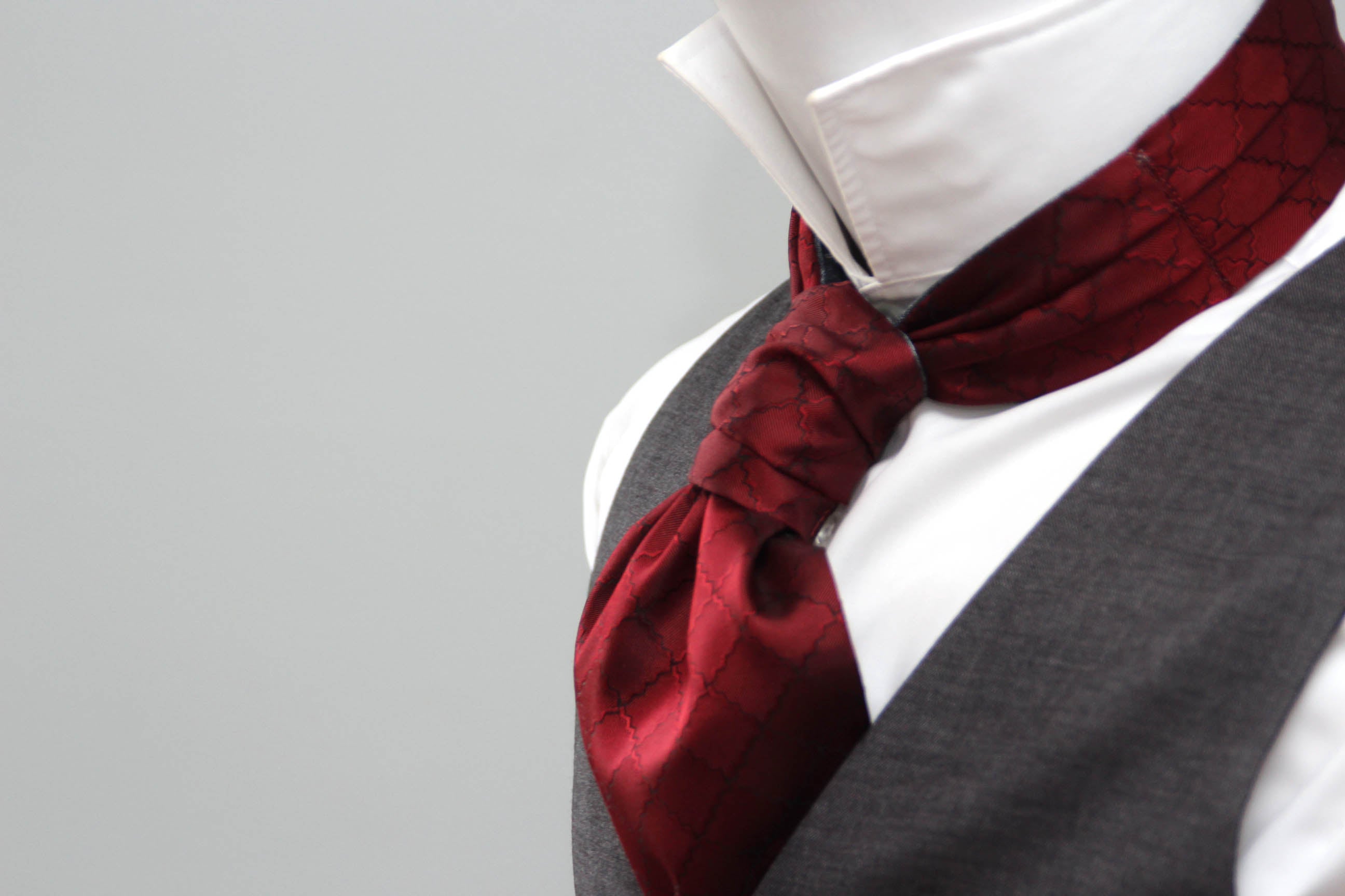 Red ascot tie on a white shirt with gray vest against a light gray background