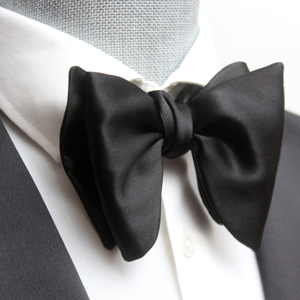 A large black silk bow tie tied around a white collar, with a smooth, satin finish and rounded ends.
