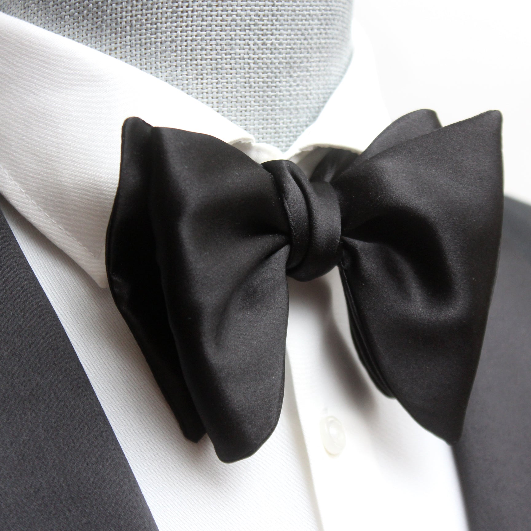 A large black silk bow tie tied around a white collar, with a smooth, satin finish and rounded ends.