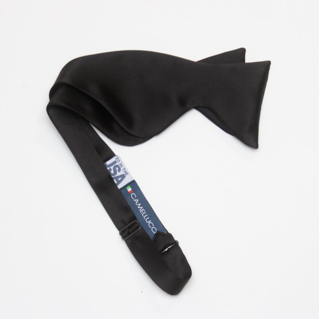 A large black silk bow tie tied around a white collar, with a smooth, satin finish and rounded ends.