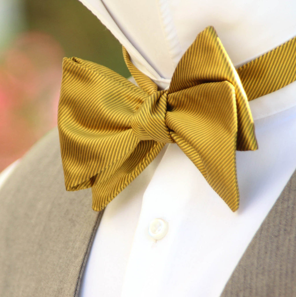 A mustard yellow self-tied bow tie on a mannequin dressed in a suit, showcasing the bow tie's color and shape against a blurred floral background.