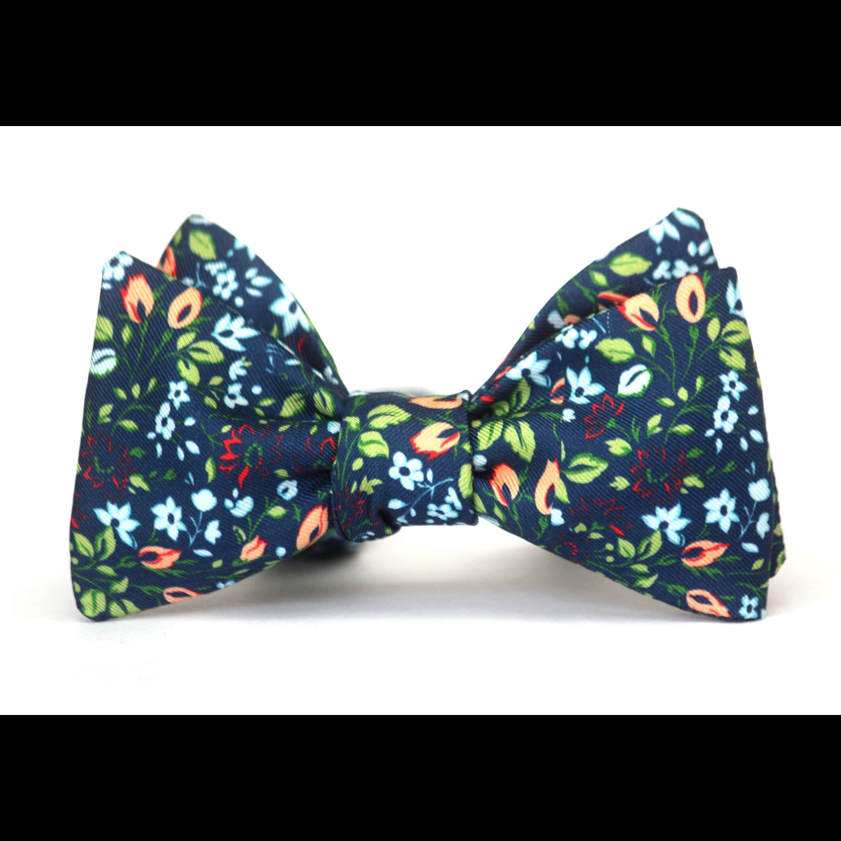A pre-tied bow tie featuring a dense floral pattern on a dark navy blue background, with green leaves, light blue and white flowers, and peach-orange accents, shown against a plain white background.