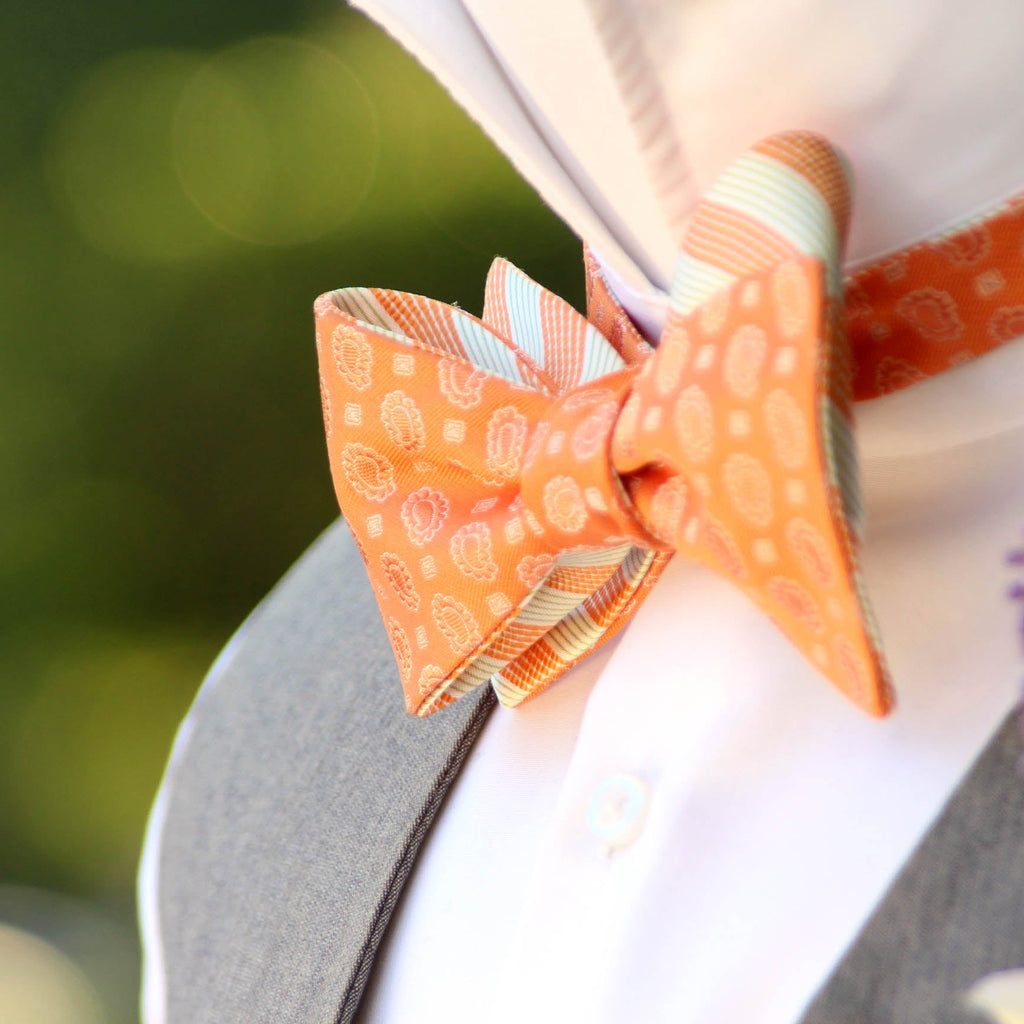 Close-up of an orange paisley stripe bow tie tied on a mannequin, with a blurred background.
