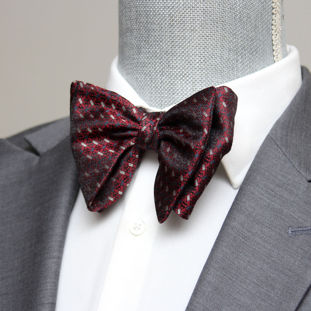 A red and black bow tie with a butterfly pattern, displayed on a mannequin wearing a grey suit and a white shirt.