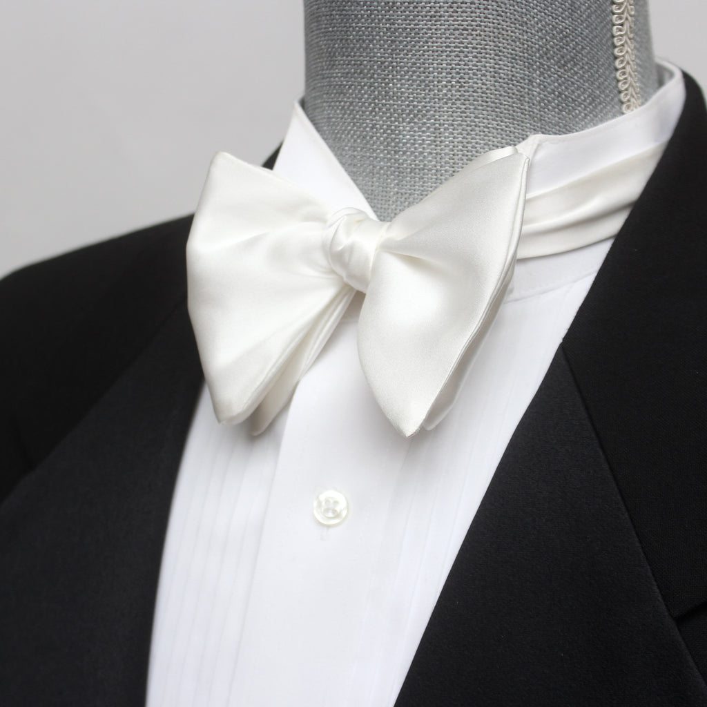 A white silk bow tie displayed on a mannequin wearing a black tuxedo and a white shirt. The bow tie has a classic butterfly shape and is attached to the mannequin's neck.
