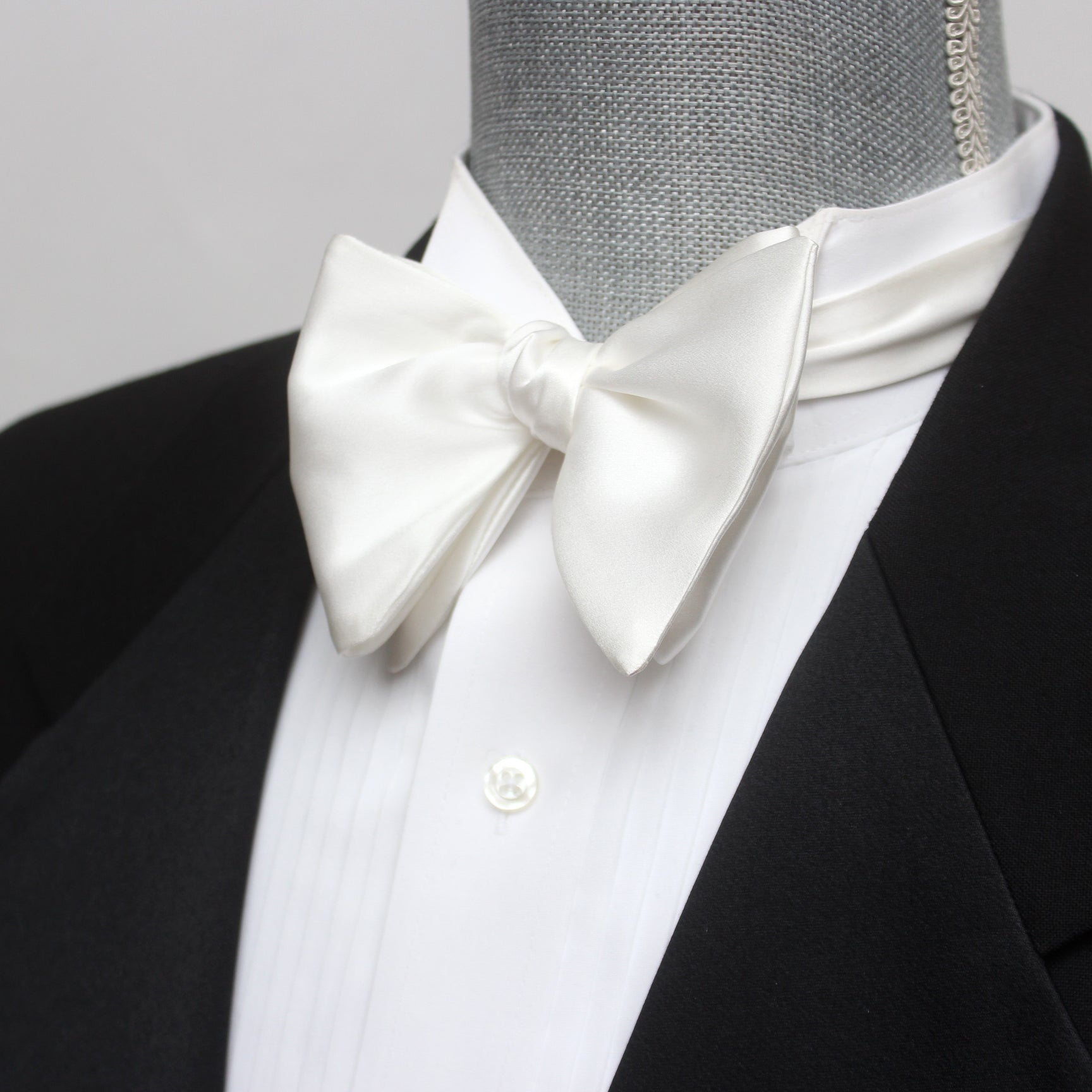 A white silk bow tie displayed on a mannequin wearing a black tuxedo and a white shirt. The bow tie has a classic butterfly shape and is attached to the mannequin's neck.